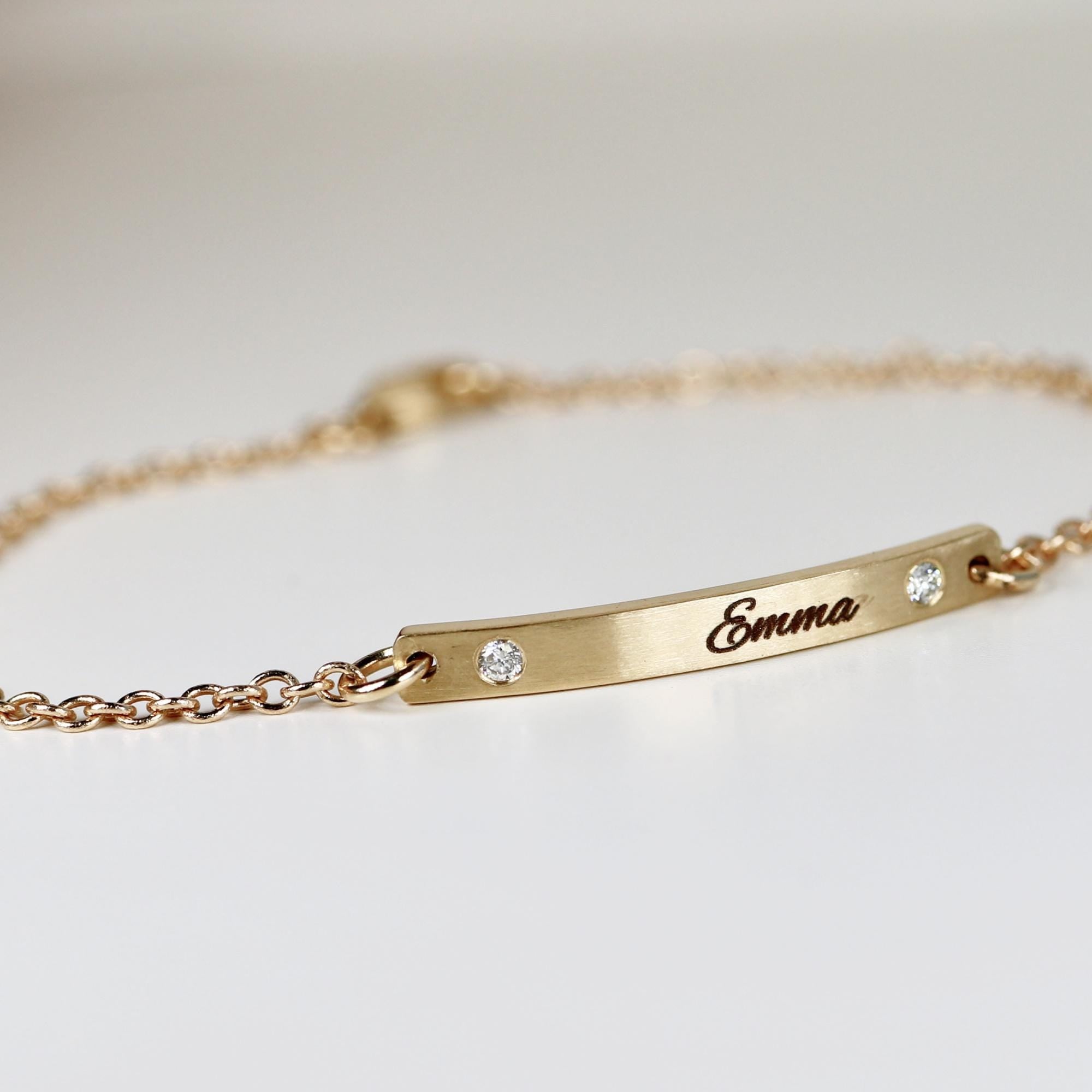 A gold bracelet with a delicate chain and a rectangular bar. The bar is engraved with the name "Emma" and features two small, sparkling gemstones. The bracelet is designed for everyday wear.