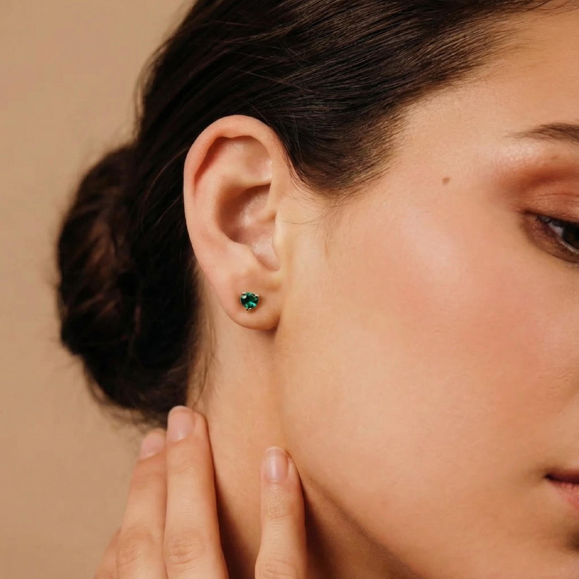 Green emerald heart earrings in solid gold, dainty gemstone studs for everyday wear, worn on ear.