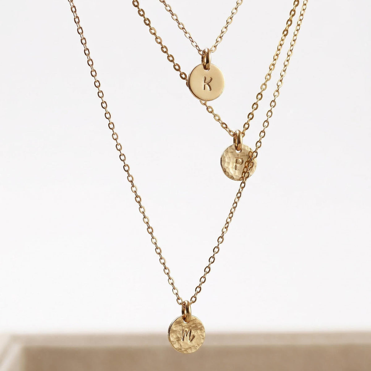 3 gold necklaces with three small, circle disc pendants. The pendants are hand stamped with the letters "M", "R", and "B".
