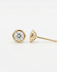 One round, faceted clear gemstone stud earring set in a gold bezel, with a second earring lying on its side showing the post.