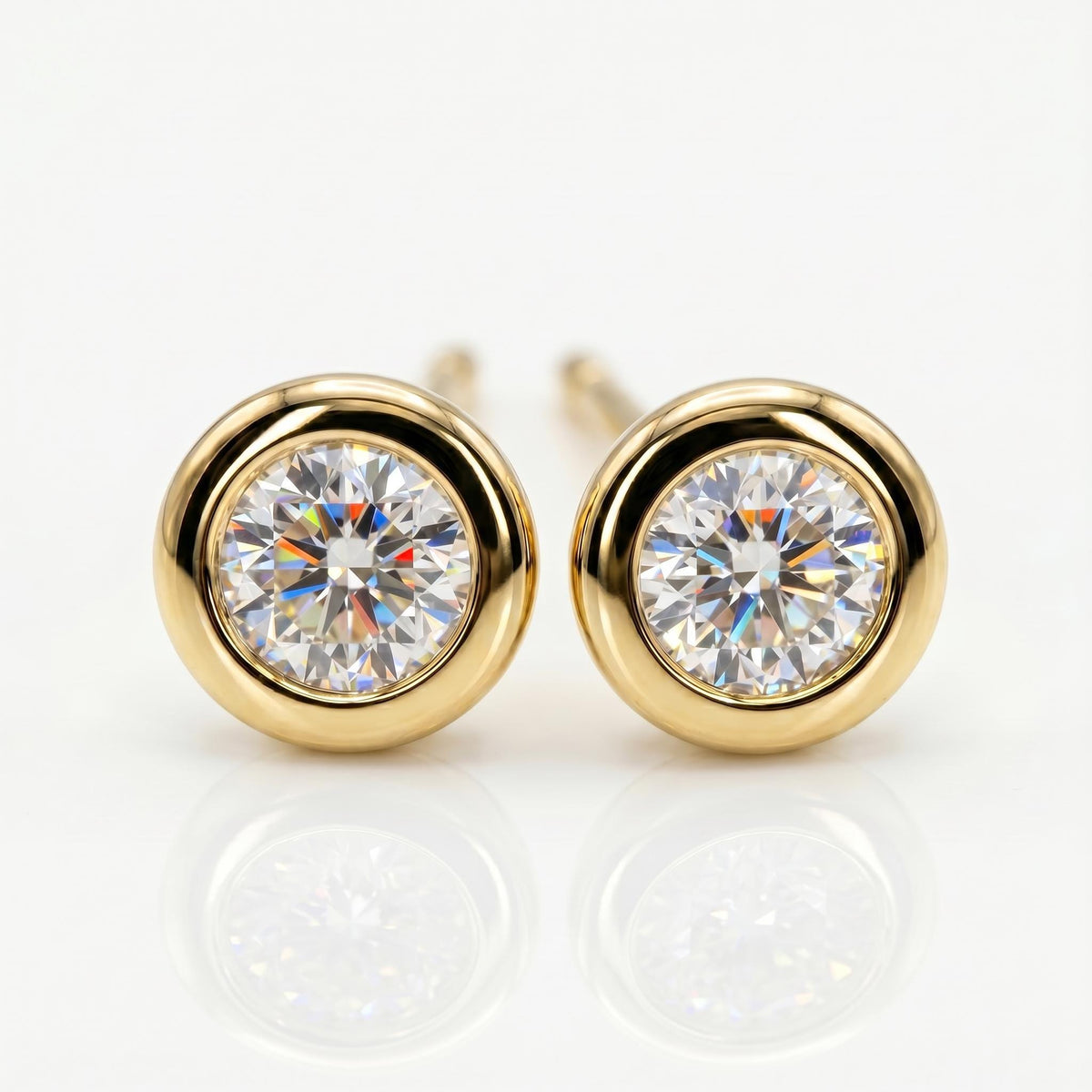 A pair of round, faceted clear gemstone stud earrings, each set in a smooth, polished gold bezel, with reflections below.