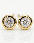 A pair of round, faceted clear gemstone stud earrings, each set in a smooth, polished gold bezel, with reflections below.