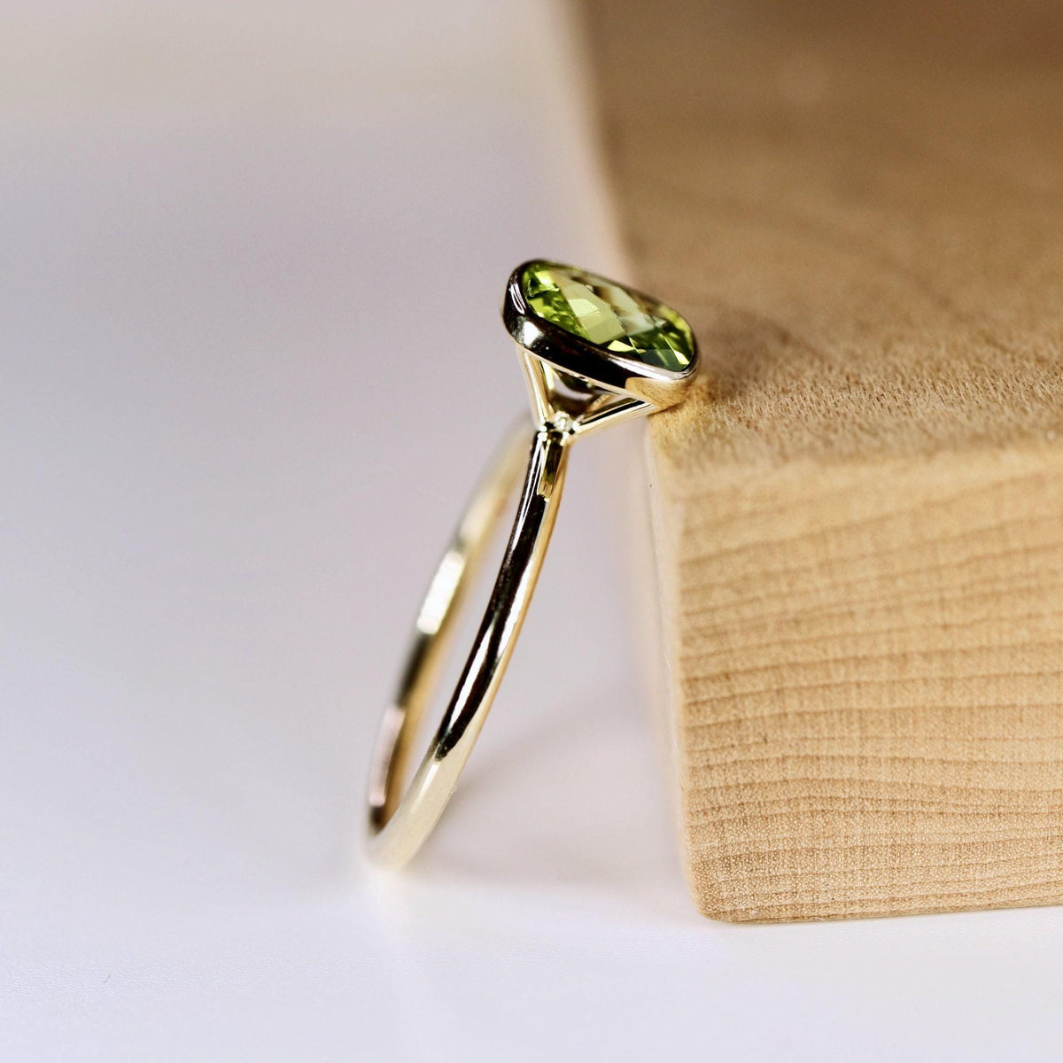 A gold ring with a bezel-set, cushion-cut green peridot gemstone stands upright against a light brown wooden block.