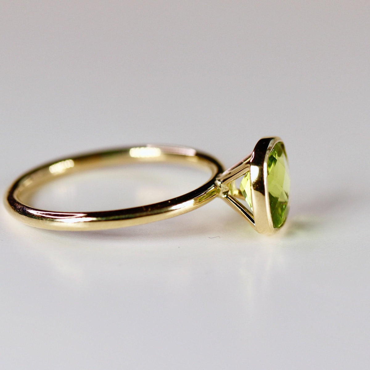 A side view of a gold ring with a bezel-set, cushion-cut green peridot gemstone on a white surface.