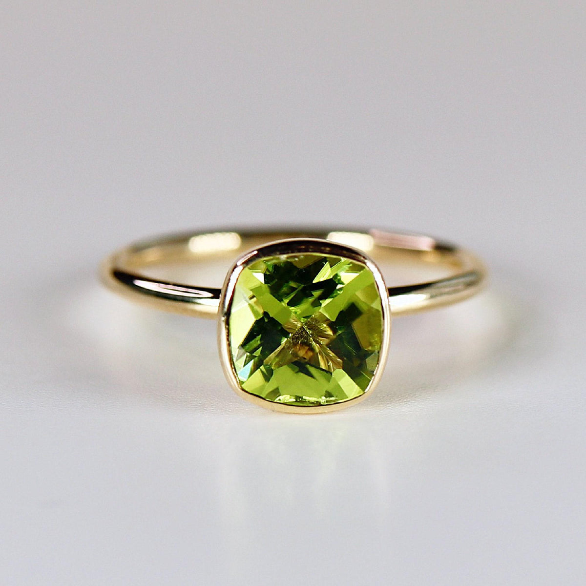 A top-down view of a gold ring with a bezel-set, cushion-cut green peridot gemstone on a white surface.