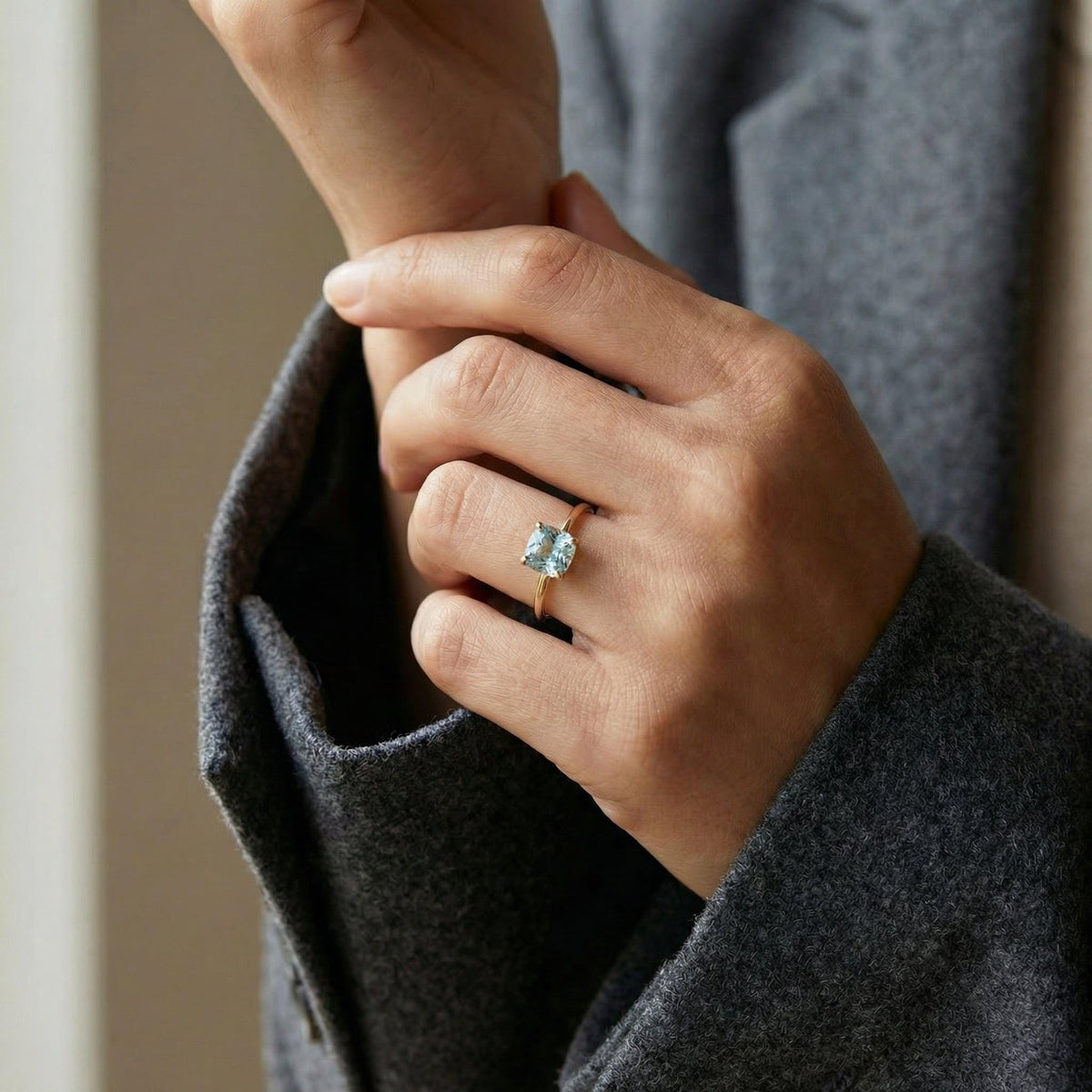 A person in a gray coat wears a gold ring with a light blue cushion-cut gemstone on their left ring finger, with their hand resting on their arm.