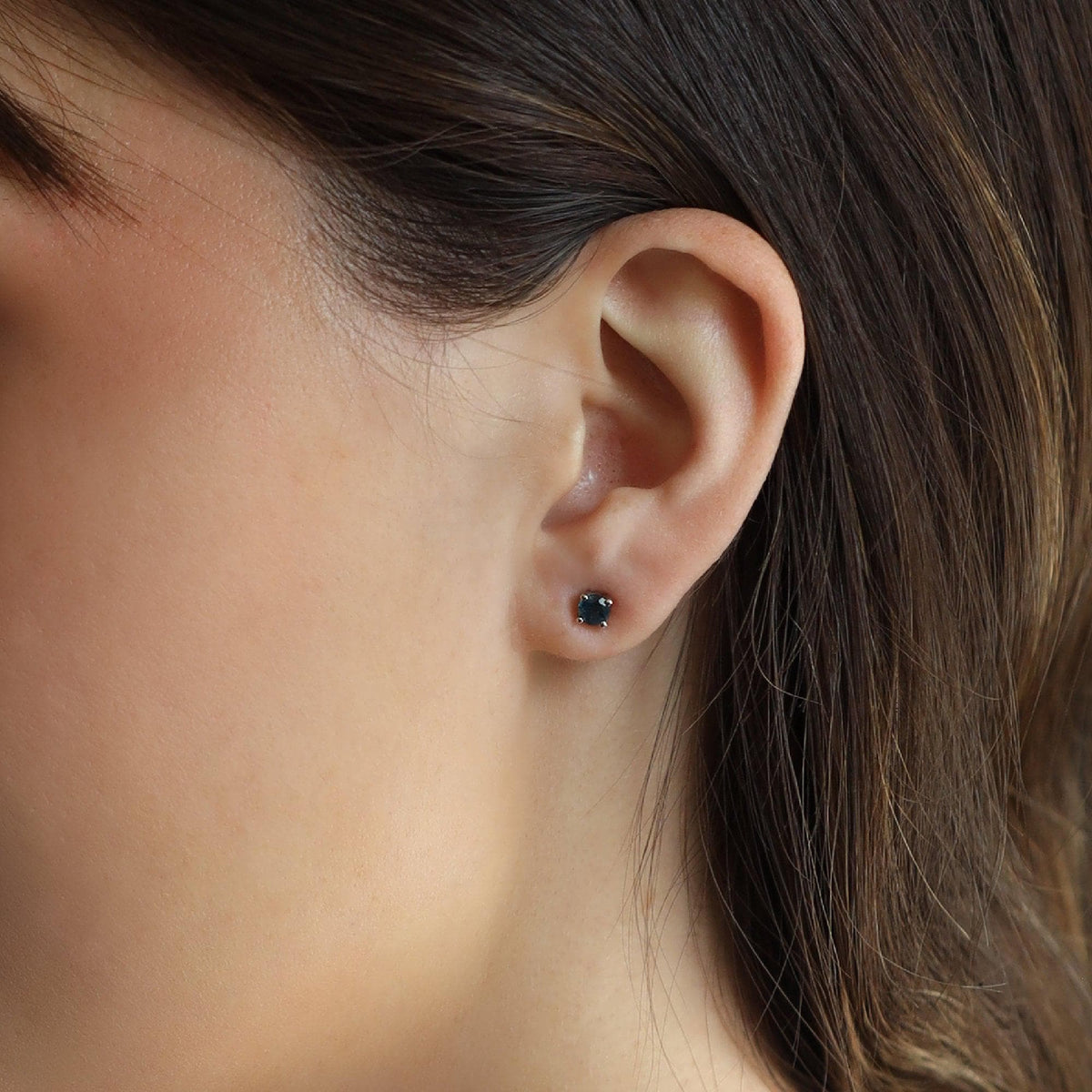 A person's ear with a single square, dark blue sapphire stud earring in a four-prong setting.