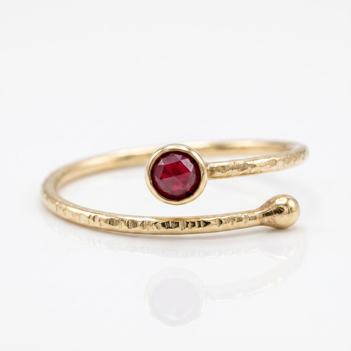 A gold bypass ring with a textured band, featuring a round ruby on one end and a small gold ball on the other, on a white surface.