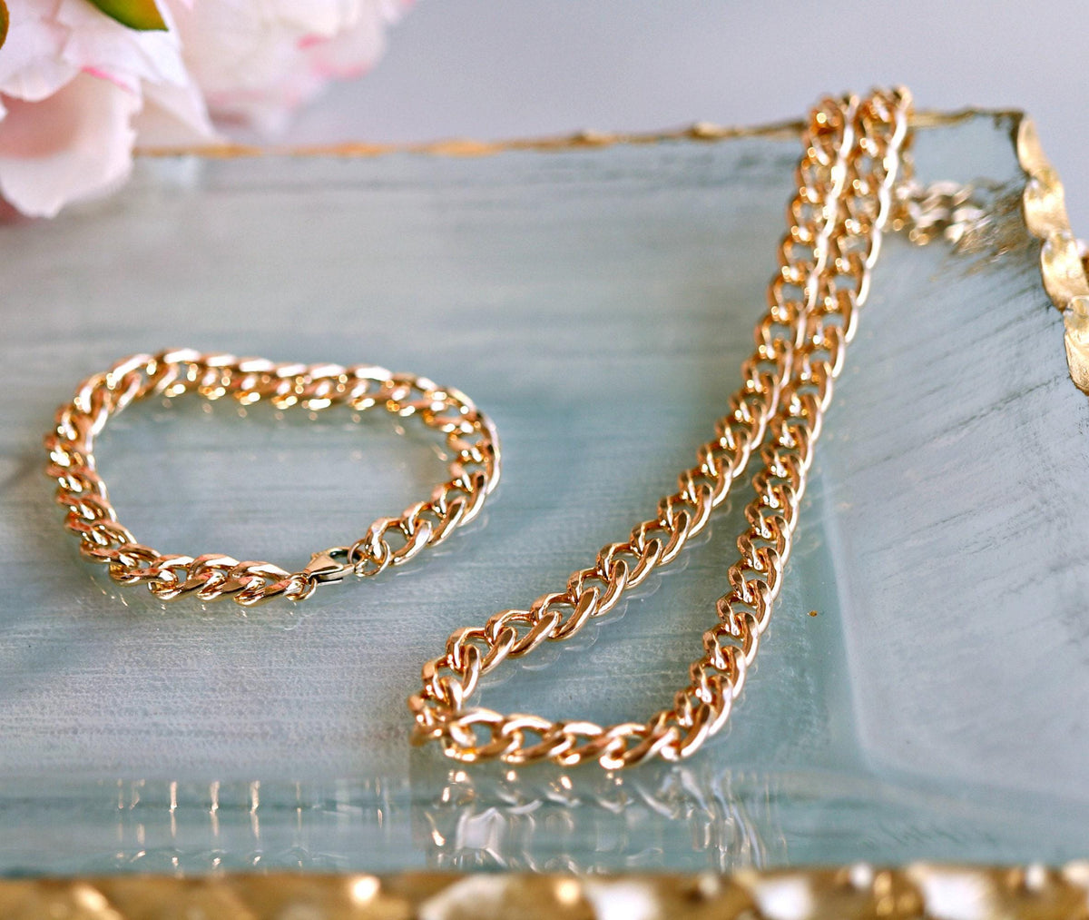A gold Cuban chain bracelet and a gold Cuban chain necklace lie on a light blue and gold tray.