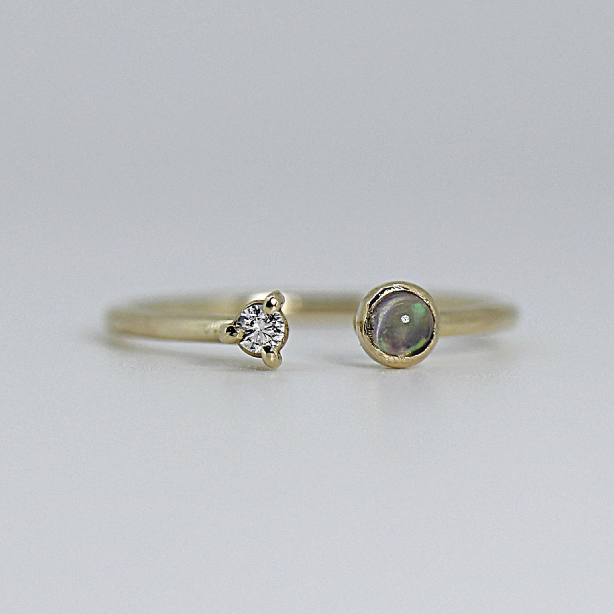 A thin gold open cuff ring with a small, clear diamond in a prong setting on the left and a round, dark iridescent opal on the right.