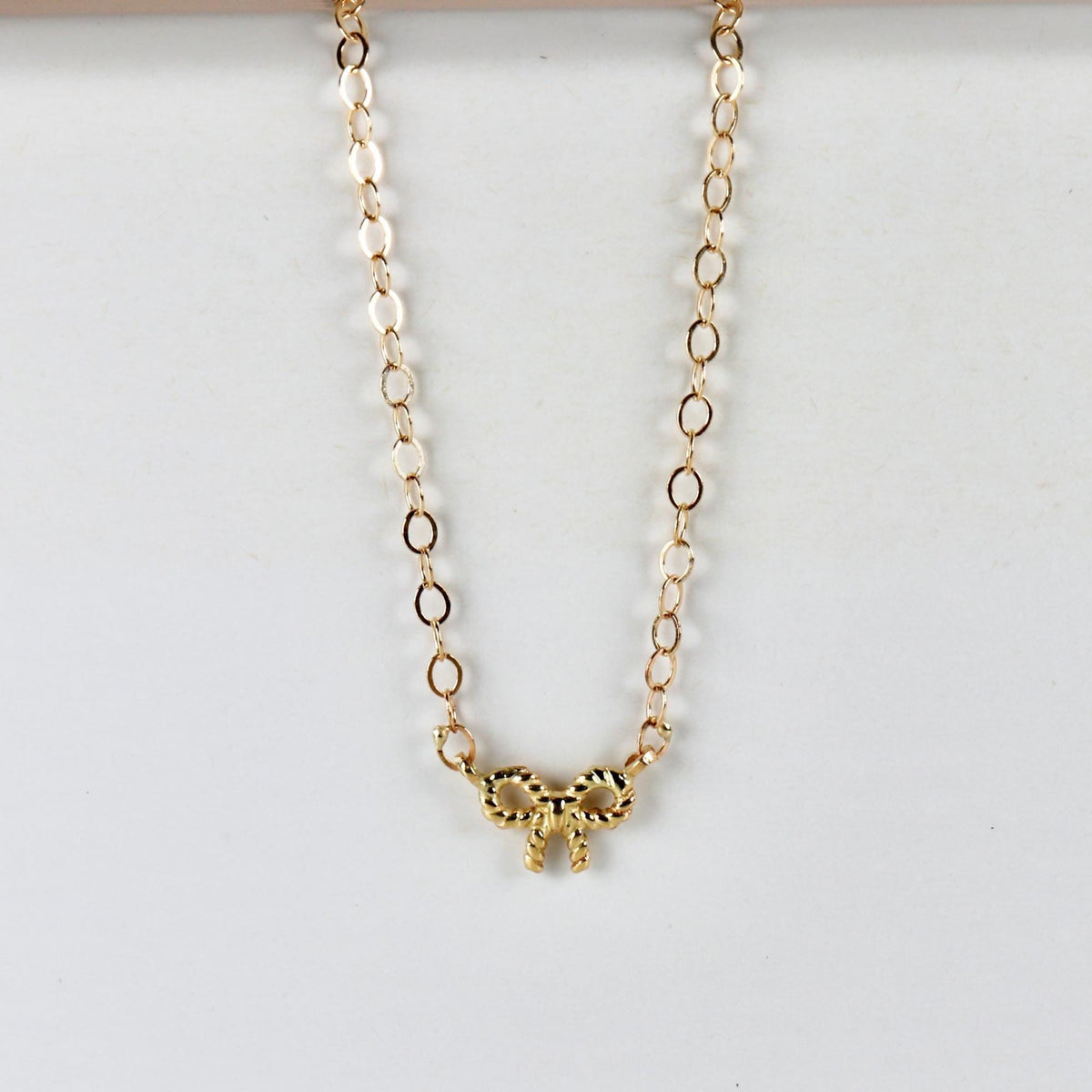 A close-up of a gold chain necklace with a twisted ribbon bow charm, displayed against a white background.