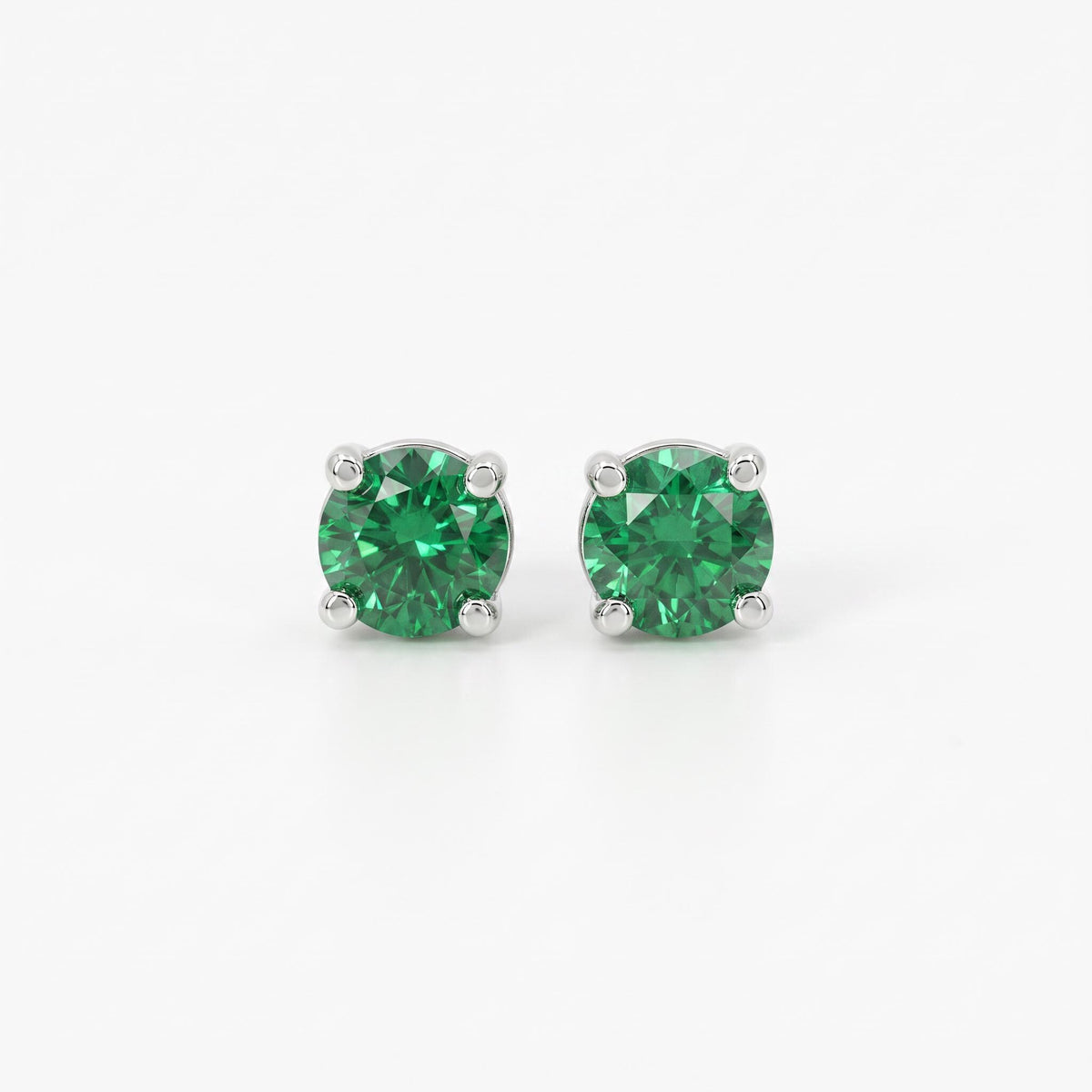 A pair of round, faceted emerald stud earrings, each set in a four-prong silver-toned setting, against a plain white background.