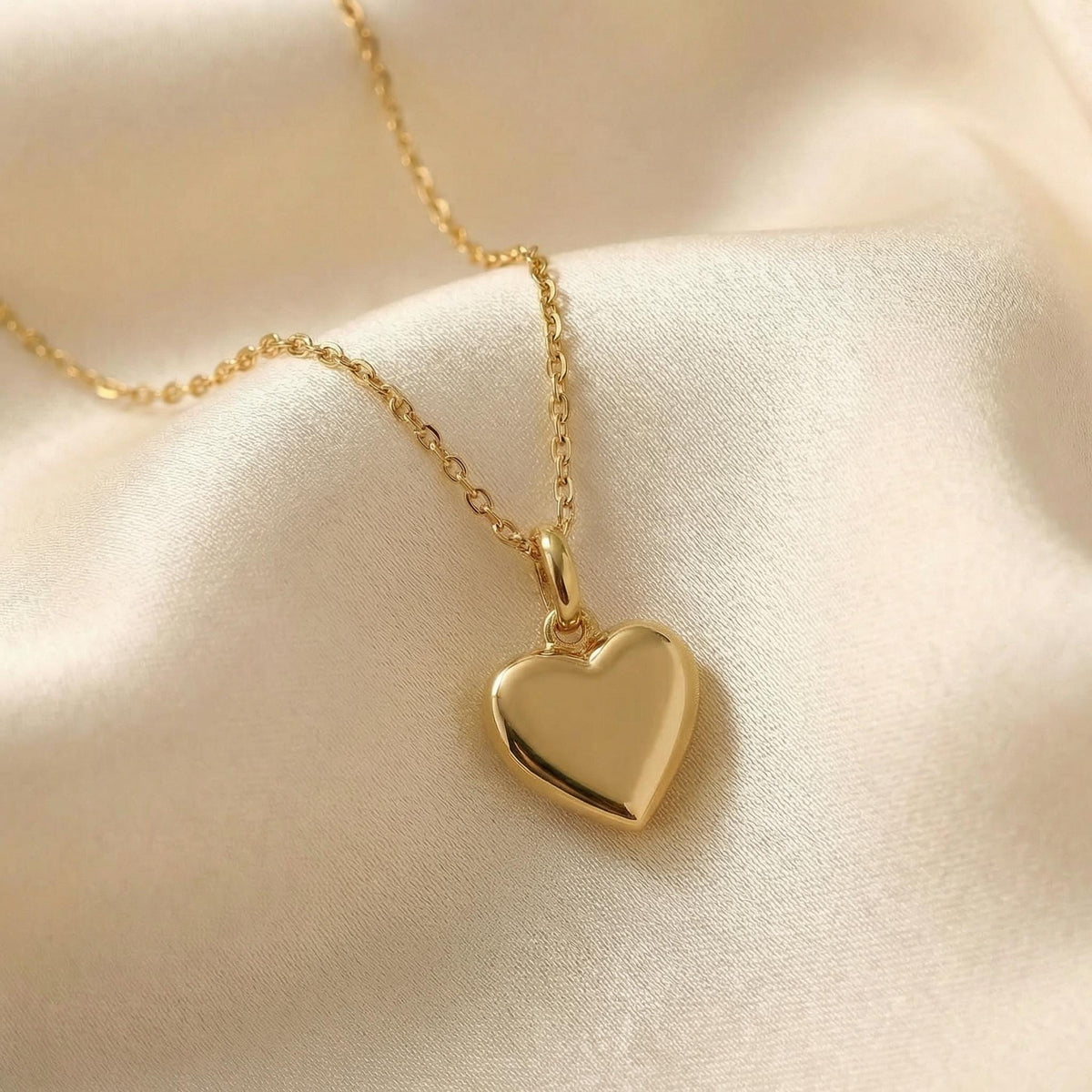 A shiny gold heart pendant hangs from a delicate gold chain, resting on a cream-colored satin fabric.