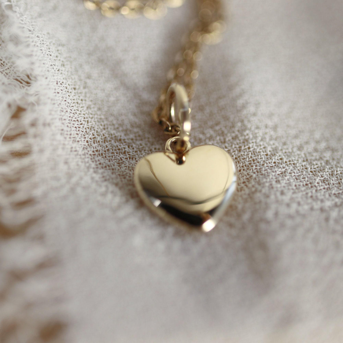 A small, shiny gold heart pendant with a bail is suspended from a delicate gold chain, resting on a textured white fabric.