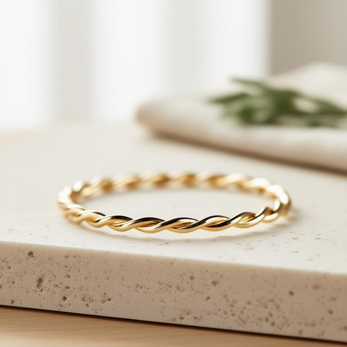 A single gold twisted rope ring rests on a light-colored stone surface, with a blurred green plant in the background.