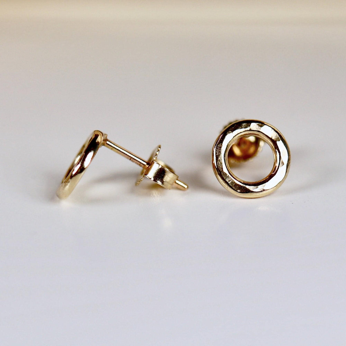 Two hammered gold open circle stud earrings are shown on a white surface, one facing forward and one angled to show its post.