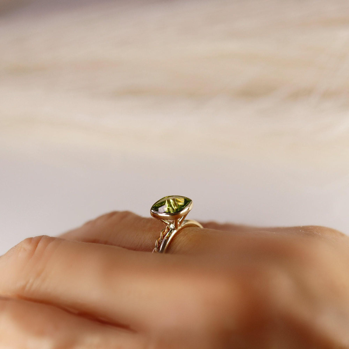A hand wears a gold ring with a cushion-cut green peridot gemstone, set in a bezel, with a blurred background.