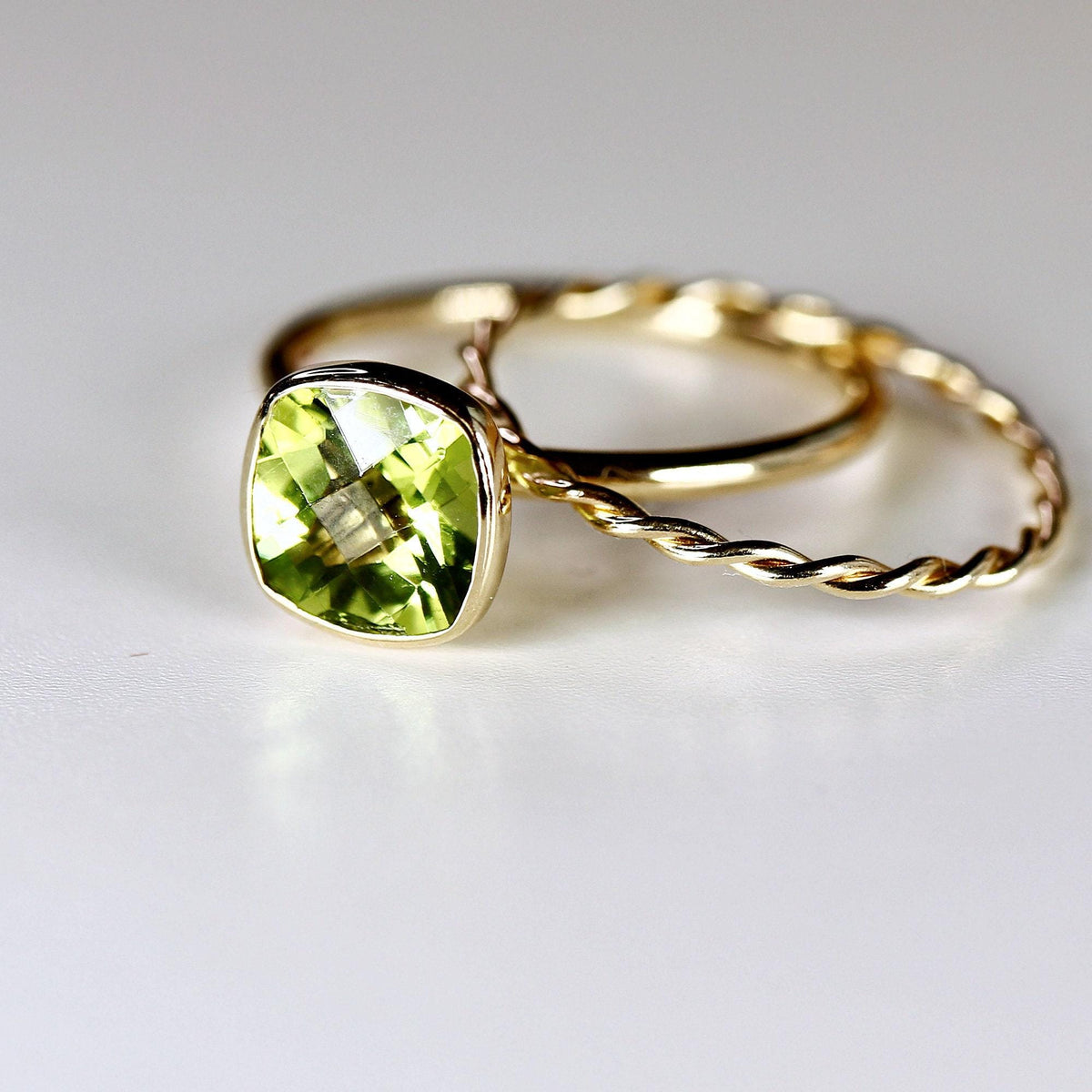 A gold ring with a cushion-cut green peridot gemstone in a bezel setting is paired with a twisted gold band on a white surface.