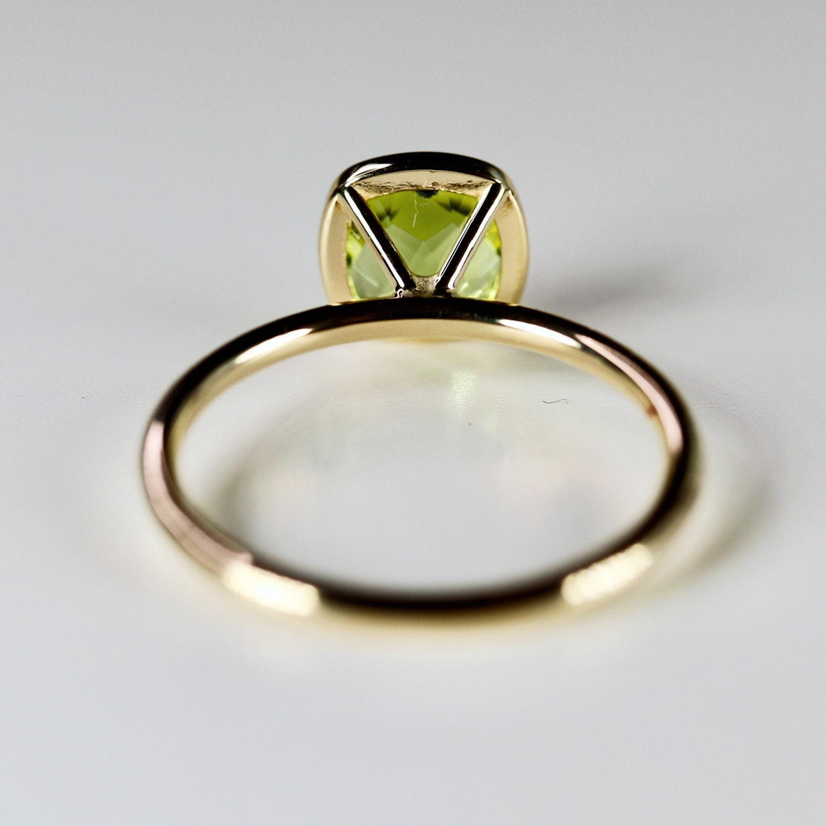 A gold ring with a bezel-set, cushion-cut green peridot gemstone is viewed from the back, showing the band and the stone&amp;