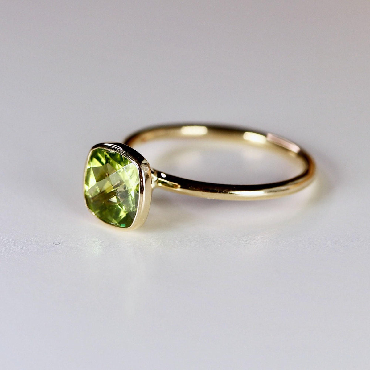 An angled view of a gold ring with a bezel-set, cushion-cut green peridot gemstone on a white surface.