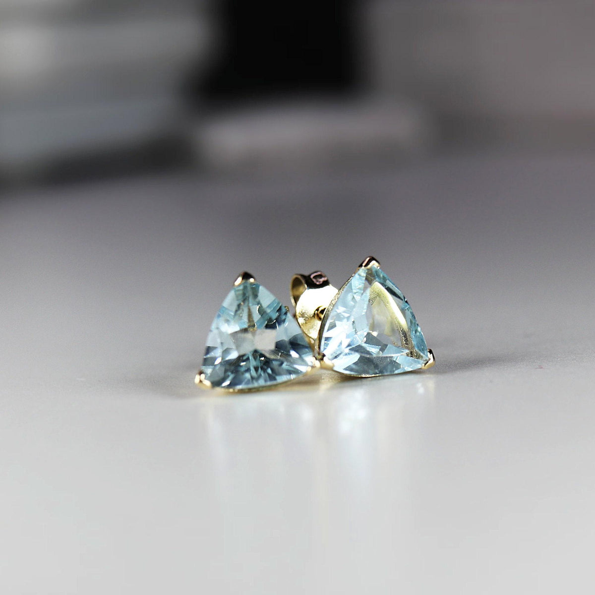 Two 14k gold stud earrings, each with a light blue trillion-cut gemstone, are angled on a white surface.