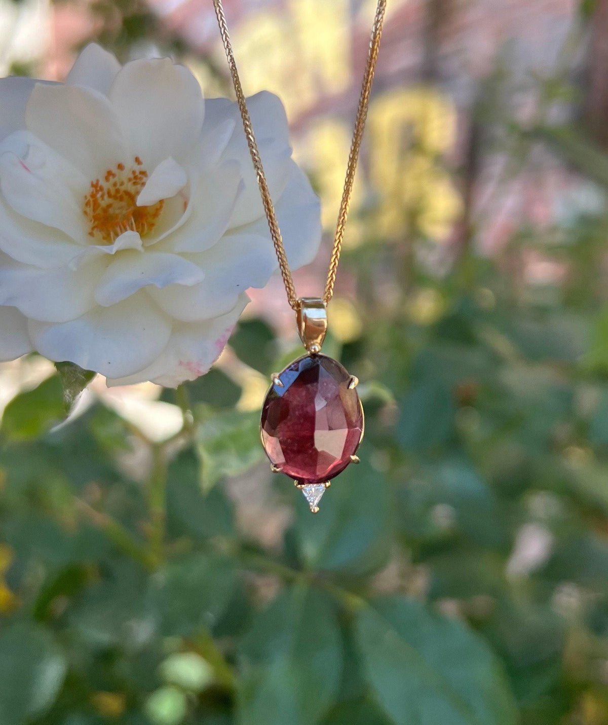 A gold necklace with an oval, faceted, dark pink tourmaline pendant, with a small triangular diamond set below it, hangs in front of a white rose and green foliage.