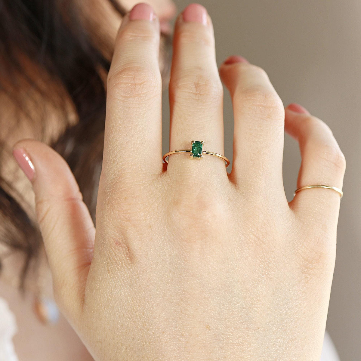 A slender gold ring with a rectangular green emerald stone on a person's ring finger, with another plain gold band on the middle finger.