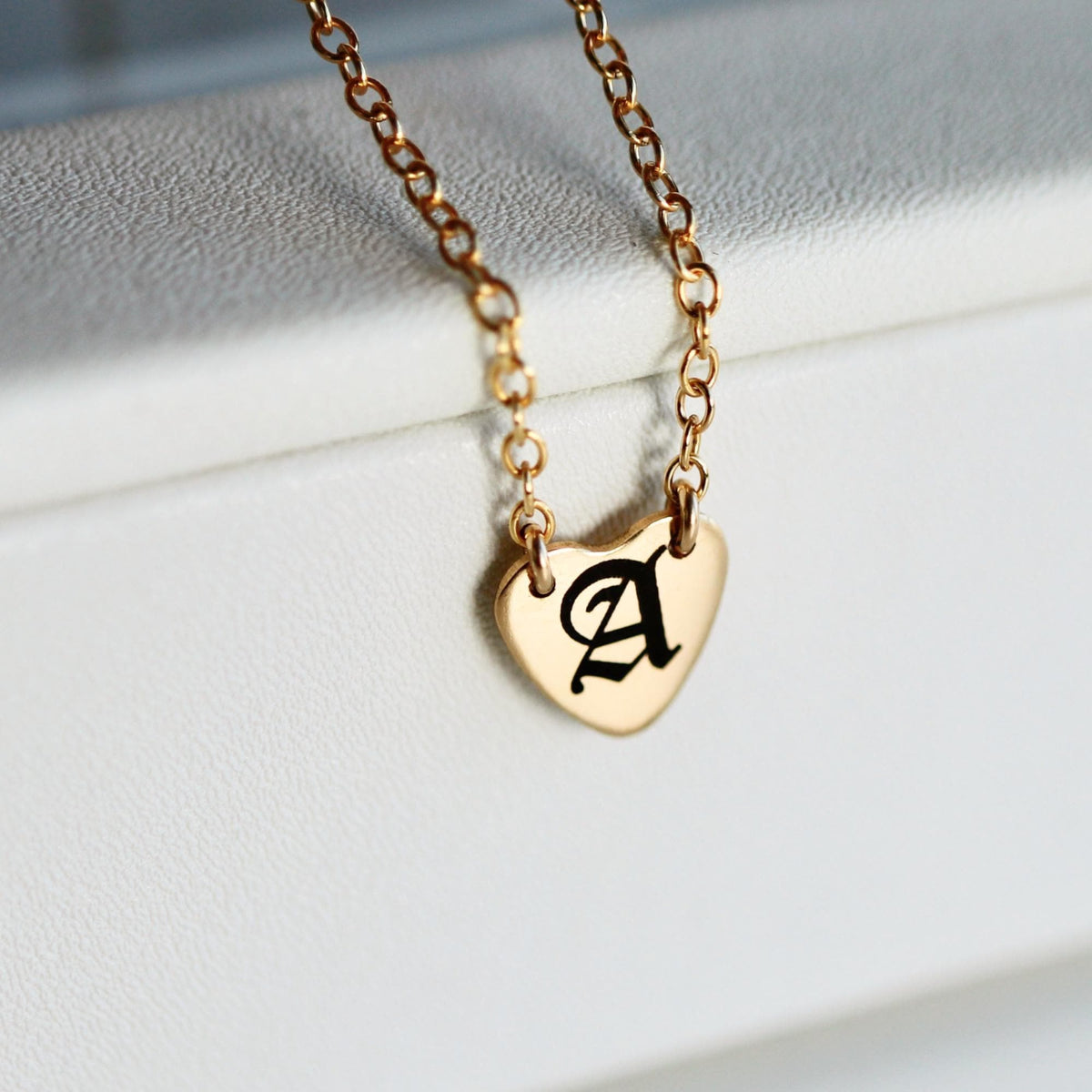 A gold chain necklace with a small heart-shaped pendant engraved with a black Gothic letter "A" rests on a white surface.