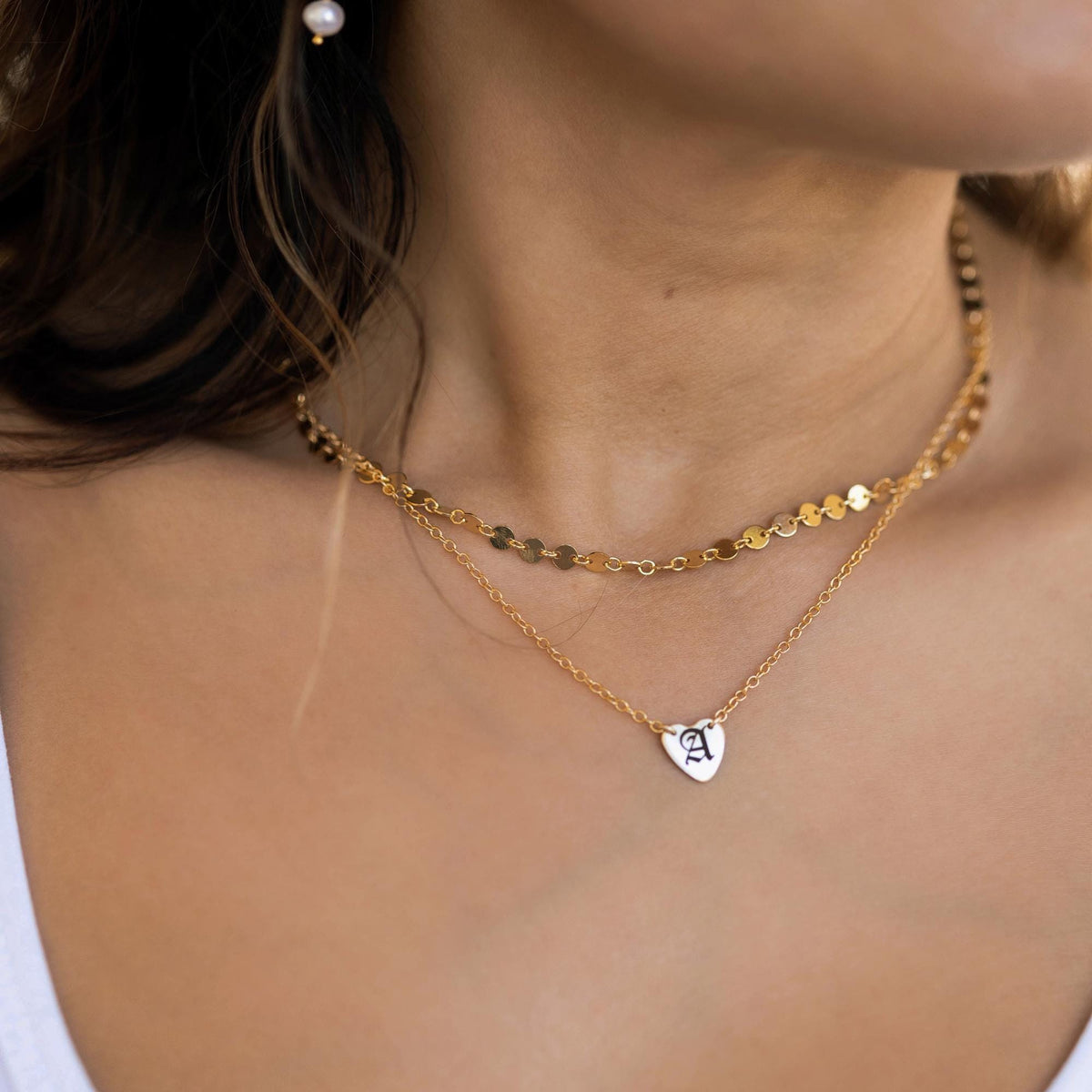 A person wears a layered gold necklace, with a choker made of flat gold discs and a longer chain with a small heart pendant engraved with a white letter &amp;quot;A.&amp;quot;