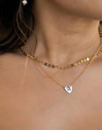A person wears a layered gold necklace, with a choker made of flat gold discs and a longer chain with a small heart pendant engraved with a white letter "A."