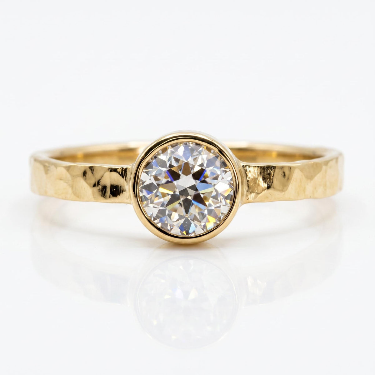 A gold ring with a hammered band and a round, bezel-set diamond is centered on a white reflective surface.