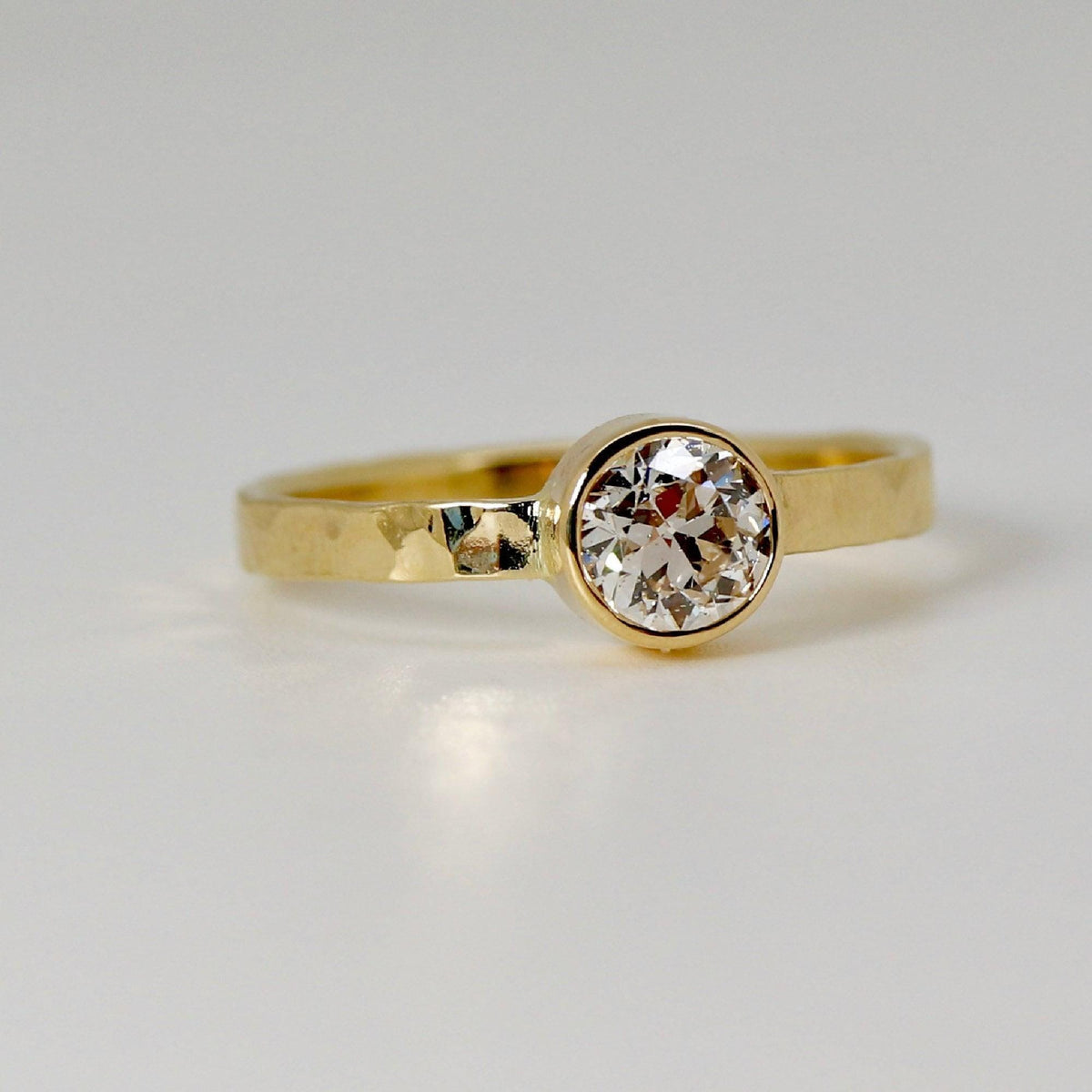 A gold ring with a hammered band and a round, bezel-set diamond is viewed from a low side angle on a white surface.