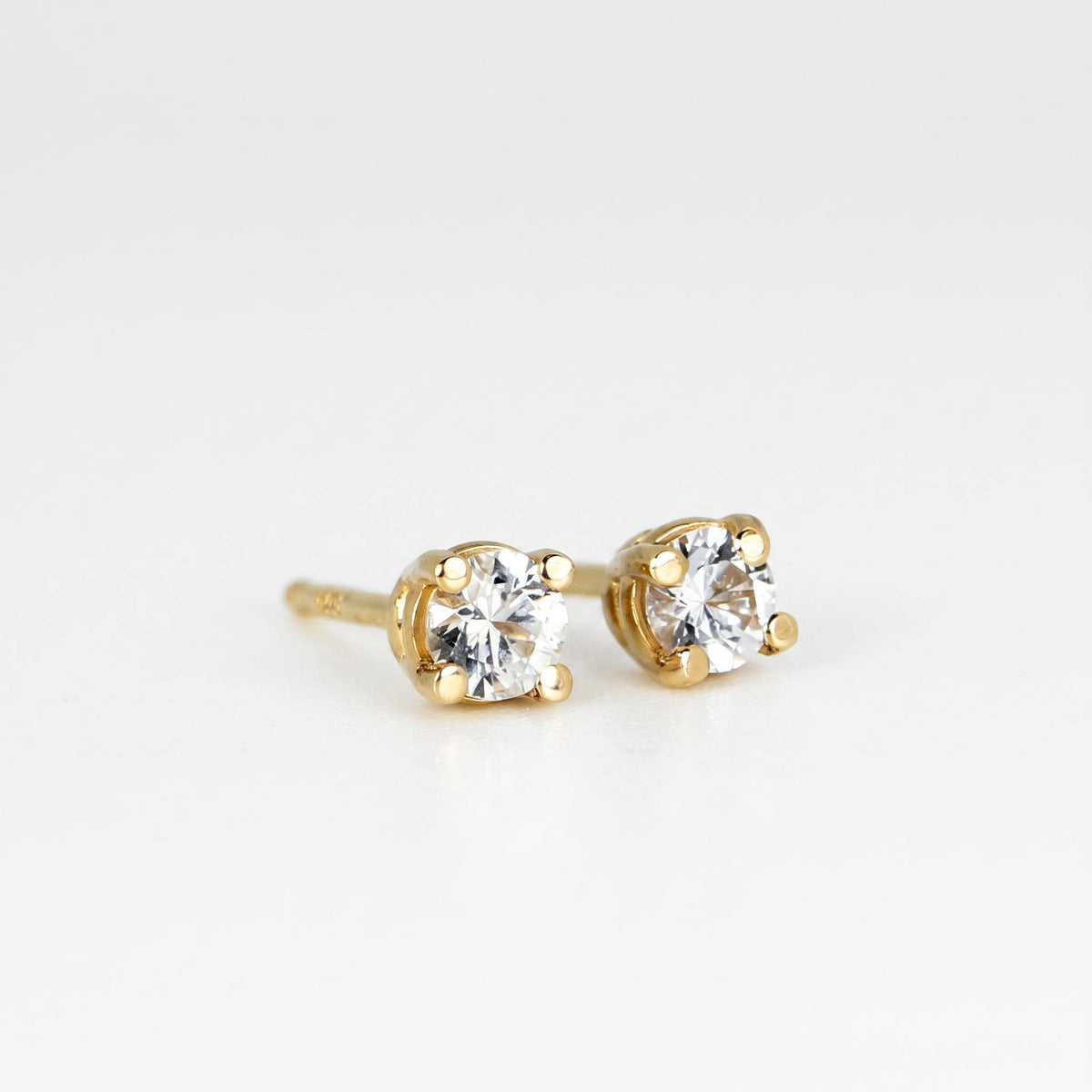 A pair of small, round, faceted white sapphire stud earrings in four-prong gold settings, viewed from above on a white background.