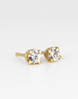 A pair of small, round, faceted white sapphire stud earrings in four-prong gold settings, viewed from above on a white background.
