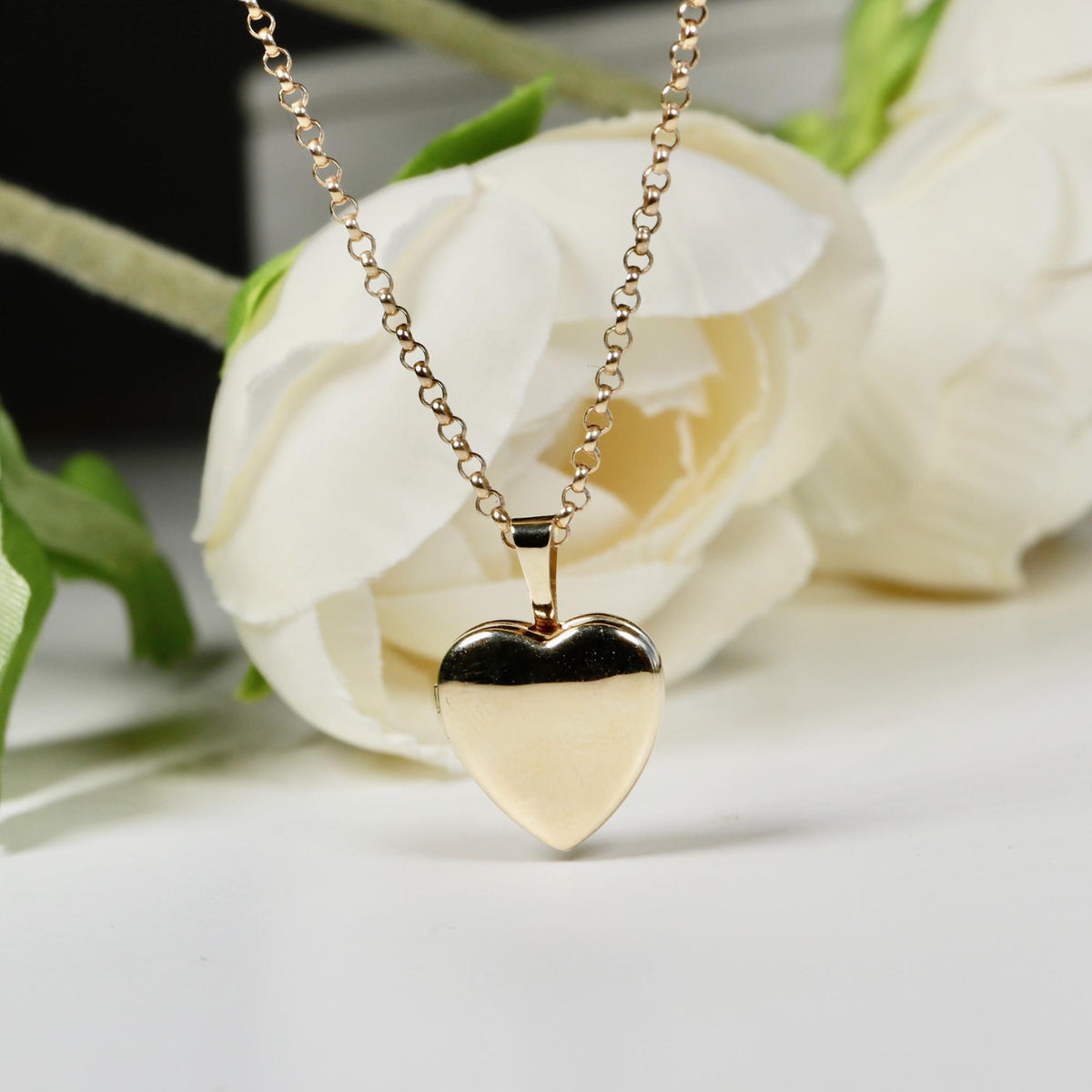 A gold heart-shaped locket on a gold chain, resting on white fabric flowers with a dark background.