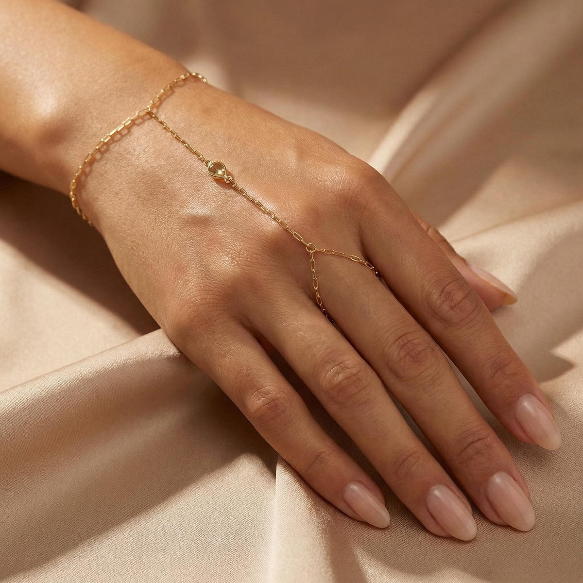 A gold hand chain with a small, light green, pear-shaped peridot gemstone is worn on a hand with manicured nails, against a light gold fabric background.