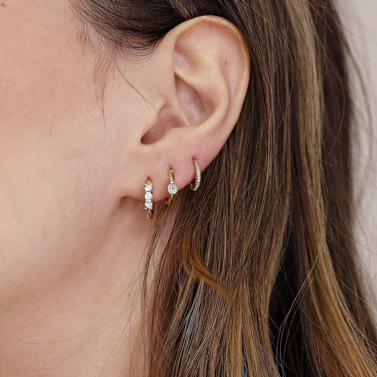 A person's ear with three gold hoop earrings: a small plain hoop, a small hoop with three white stones, and a small hoop with a single white stone.