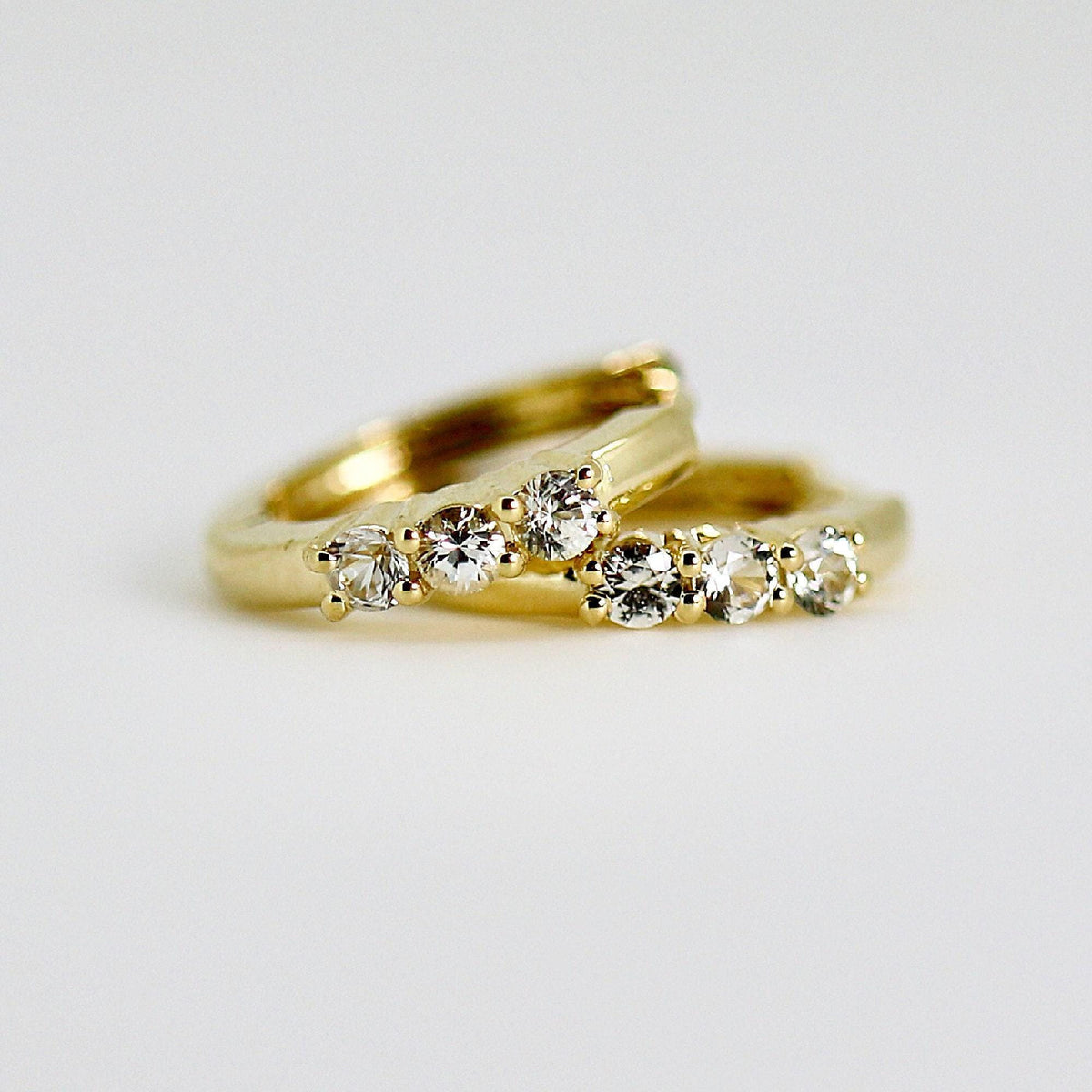 Two small gold huggie hoop earrings, each with three clear round stones set into the front, on a white background.