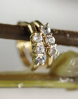 Two small gold huggie hoop earrings, each with three clear round stones, hanging from a dark brown branch.