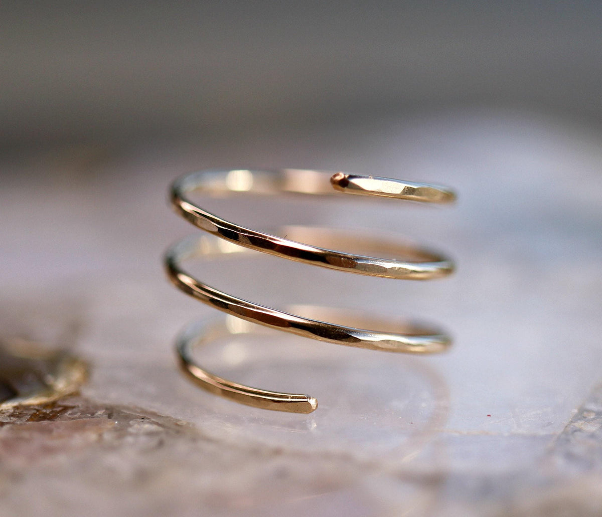 A gold-colored, three-coil spiral ring with hammered texture is shown on a blurred light-colored surface.