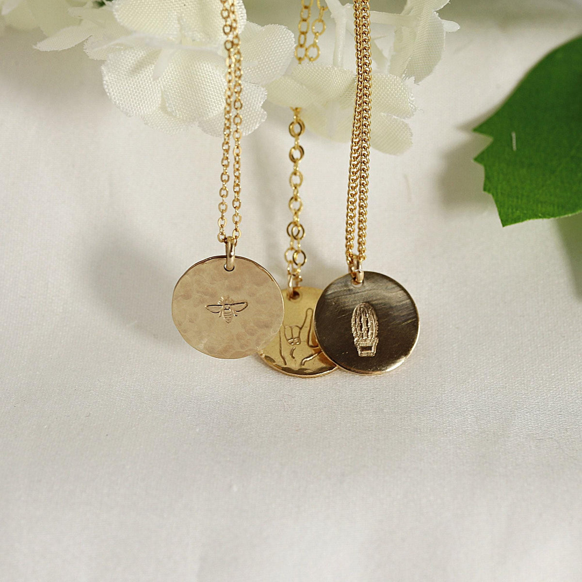 Three gold-colored necklaces hang from white flowers, each with a round pendant engraved with a bee, a hand making the "I love you" sign, or a potted cactus.