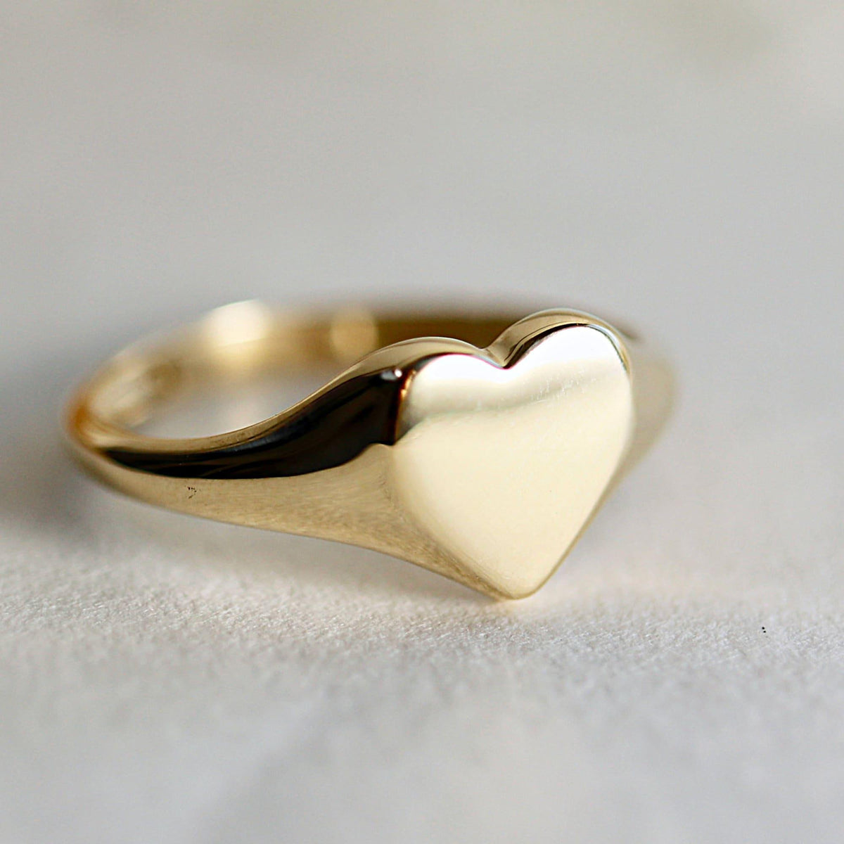 A close-up of a shiny, unengraved gold heart-shaped signet ring on a white surface.