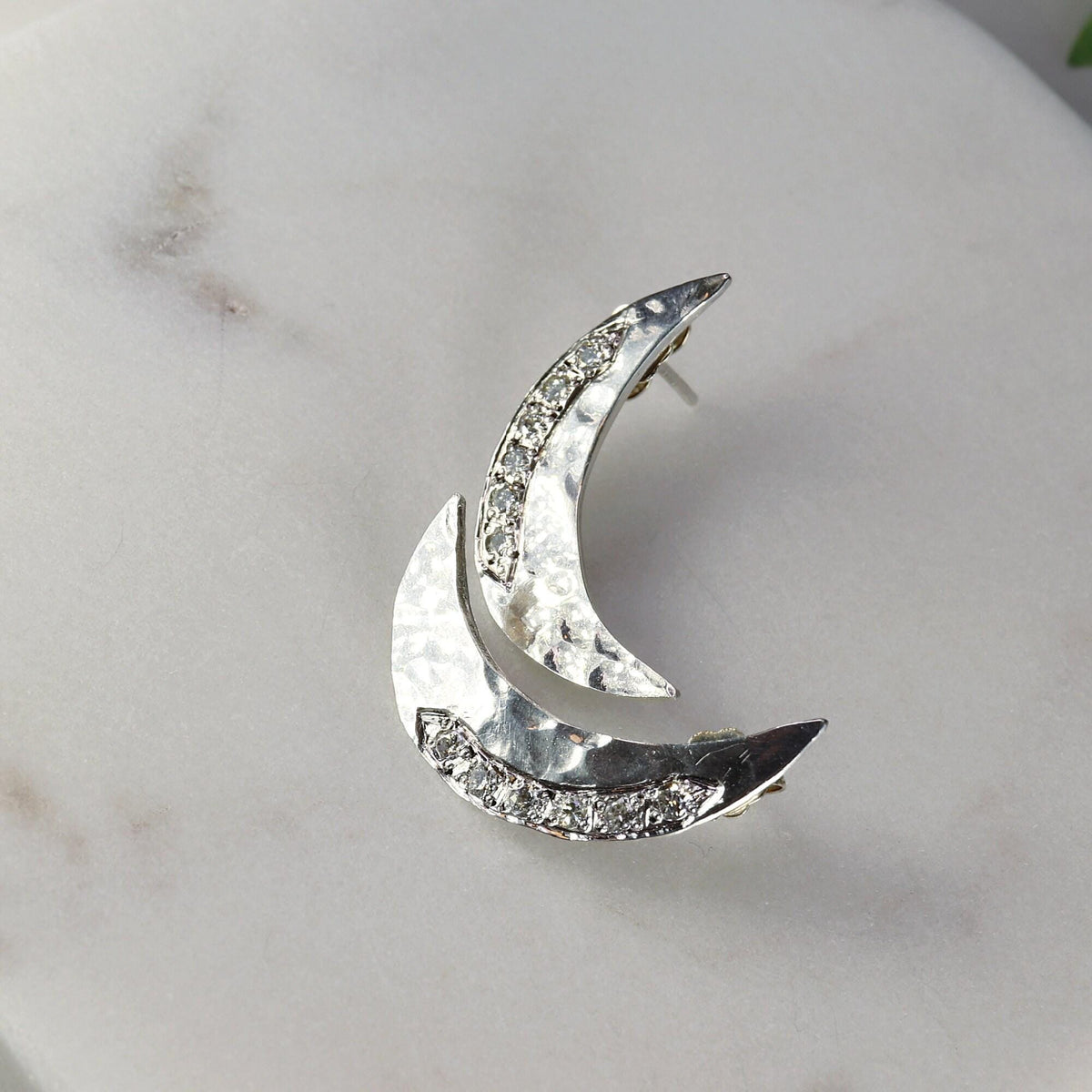 Two hammered silver crescent moon earrings with pave stones on a white marble surface.