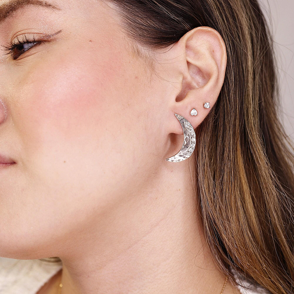 A person wearing a hammered silver crescent moon earring with small stones on the earlobe.