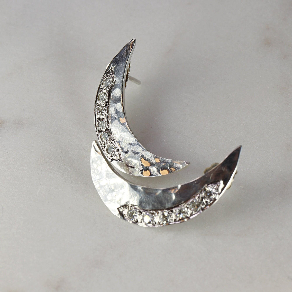 Close-up of two silver crescent moon earrings with hammered texture and embedded stones resting on a light marble background.