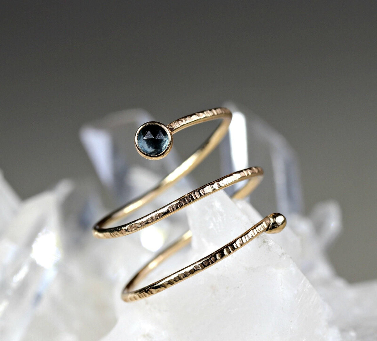 A close-up shot of a textured gold spiral ring featuring a single dark blue gemstone on a crystal background.