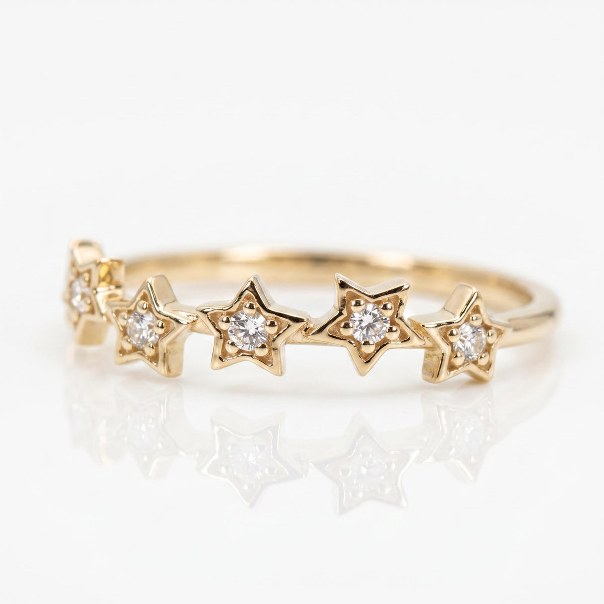 A gold ring with five diamond-studded stars across the top, reflecting on a white surface.