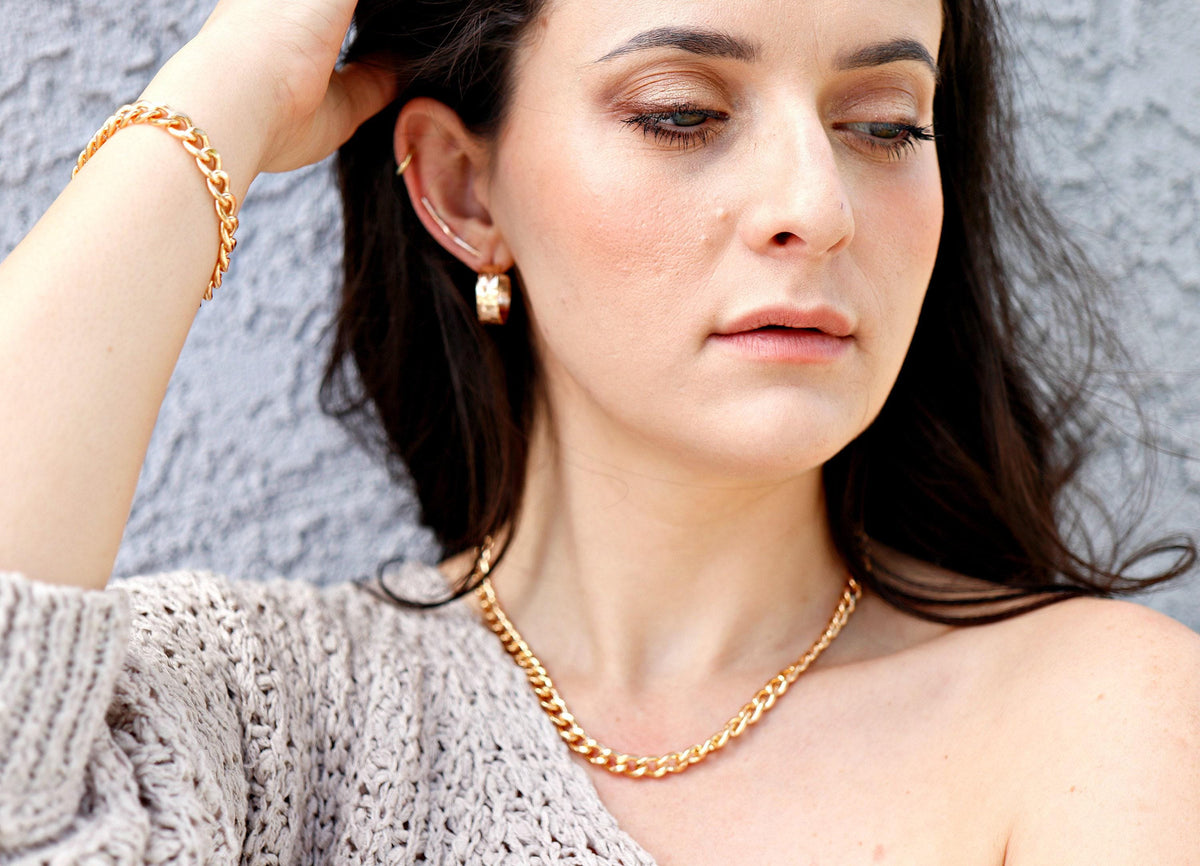A person wearing a light brown knit sweater, a gold Cuban chain necklace, and a matching gold Cuban chain bracelet.