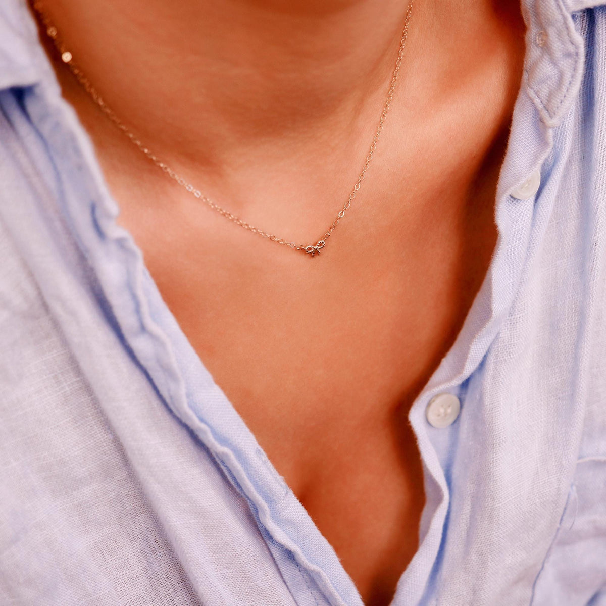 A gold chain necklace with a small bow charm is worn by a person in a light blue shirt, with the charm positioned centrally on the chest.