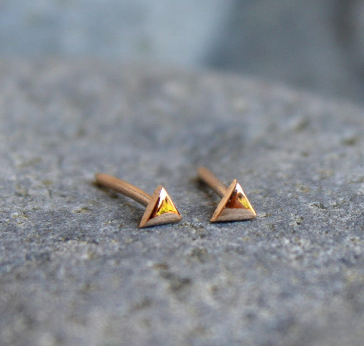 Two rose gold triangle stud earrings with posts, resting on a gray textured surface.